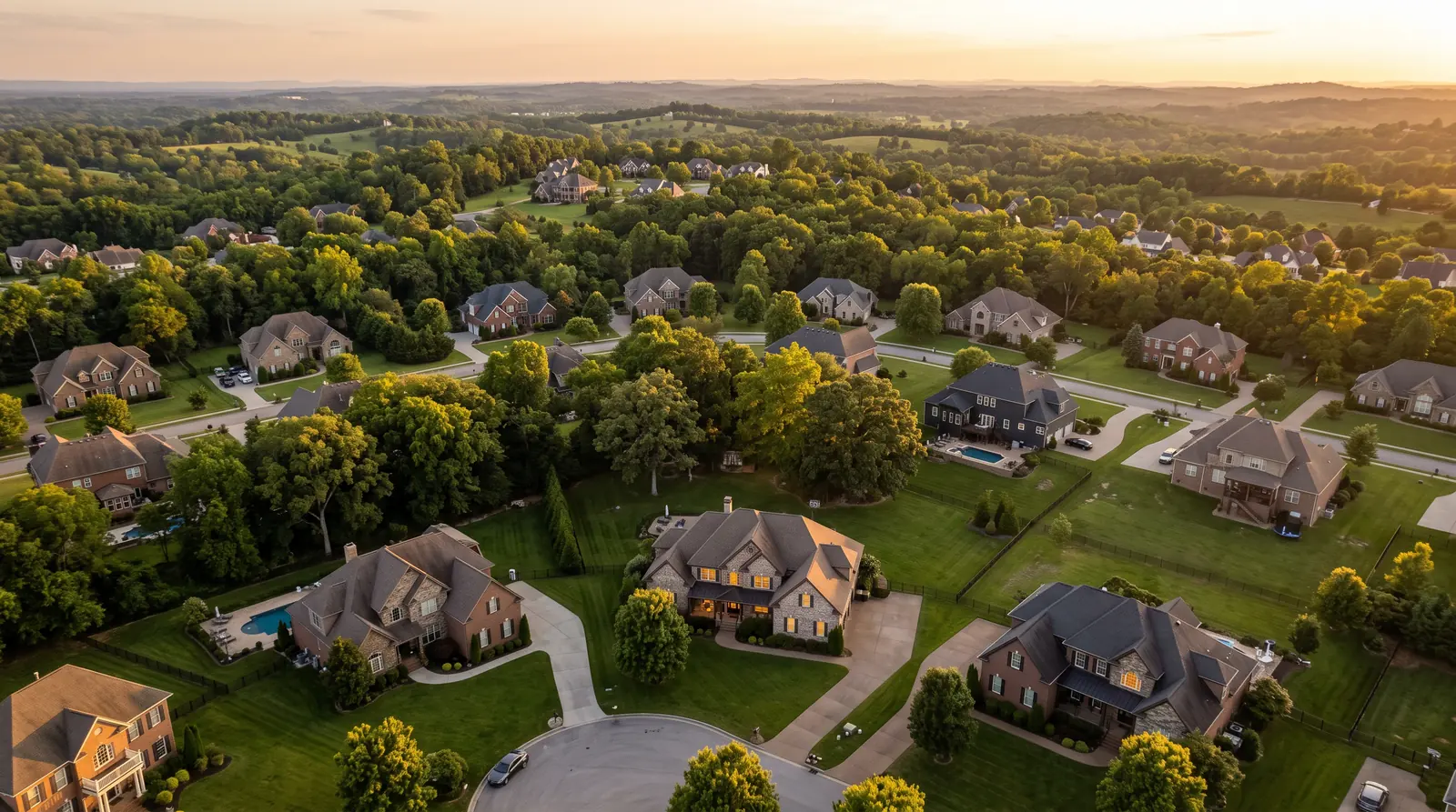 An aerial view over a green Middle Tennessee neighborhood at golden hour showing brick and stone homes, mature trees and rolling hills in the distance, with warm lights glowing in many windows