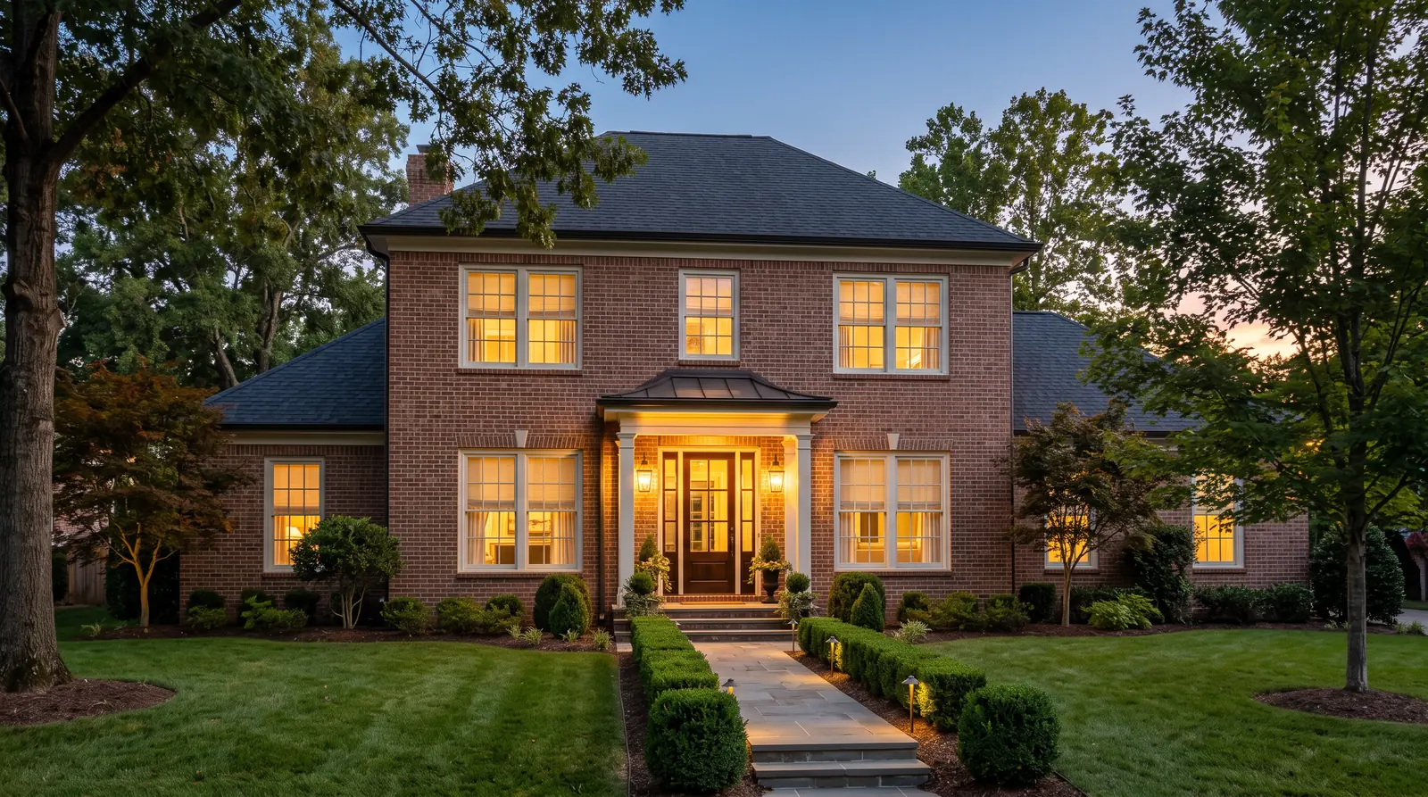 A handsome brick Tennessee home at dusk with warm light glowing through the front windows, where lowered cream shades and drapery are softly visible, trimmed hedges, a lit front door and a calm blue sky