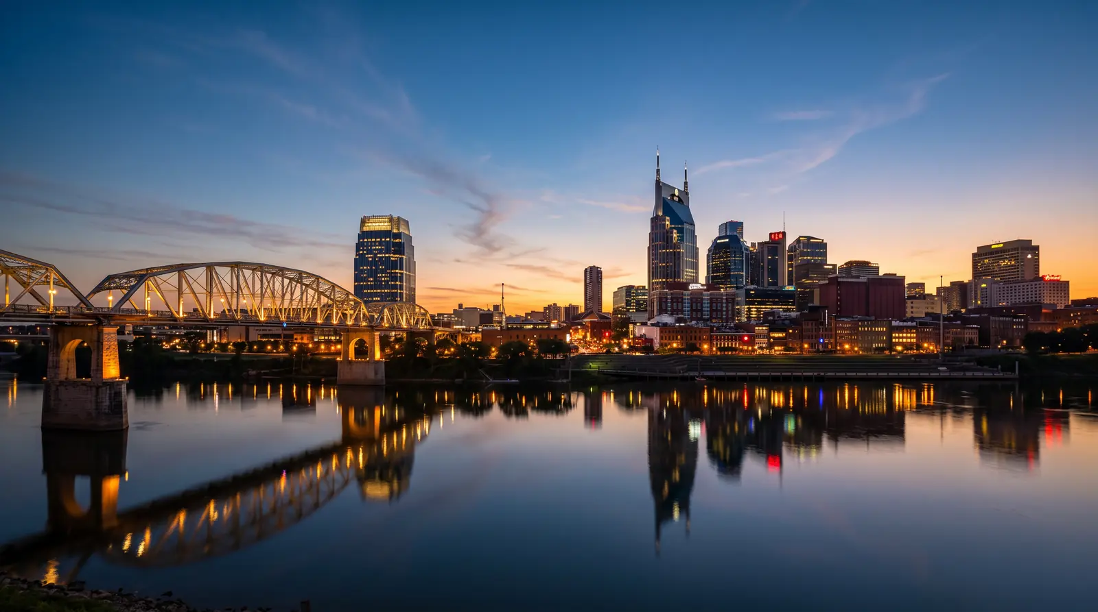 Nashville Skyline at Blue Hour | Music City Blinds The Nashville skyline glowing at blue-hour dusk beyond the Cumberland River with a calm reflection, a warm sunset gradient in the sky and the lit pedestrian bridge in the foreground