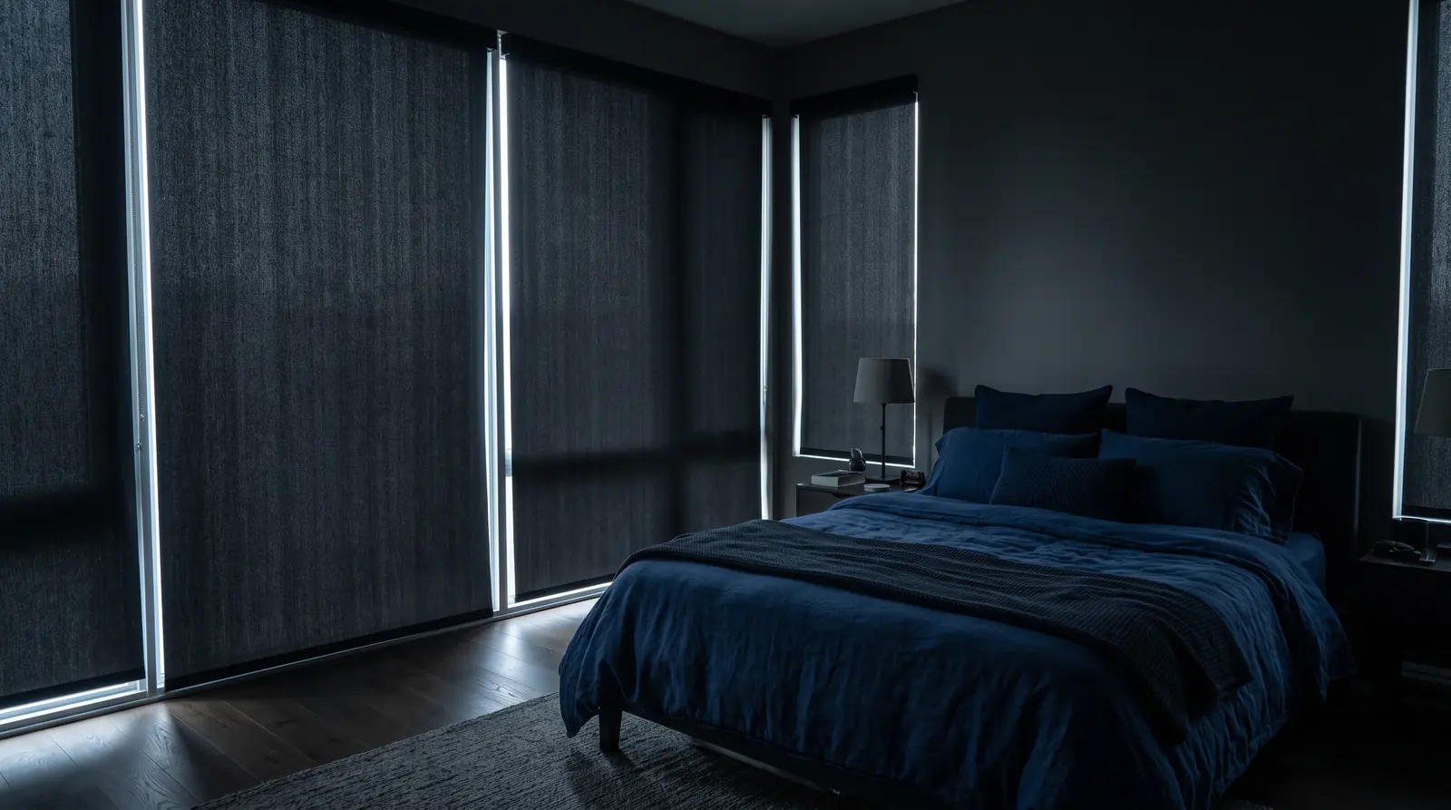 Blackout Shades in a Dark Bedroom | Music City Blinds A bedroom with tall windows dressed in thick charcoal blackout shades fully lowered with a thin edge of daylight, a bed with navy bedding, calm and dim