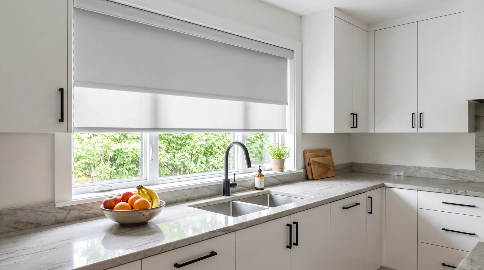 Clean Roller Shades in a Modern Kitchen | Music City Blinds A modern kitchen with a large window over a sink dressed in clean light-gray roller shades lowered halfway, white cabinets, a stone countertop and a bowl of fruit, bright daylight